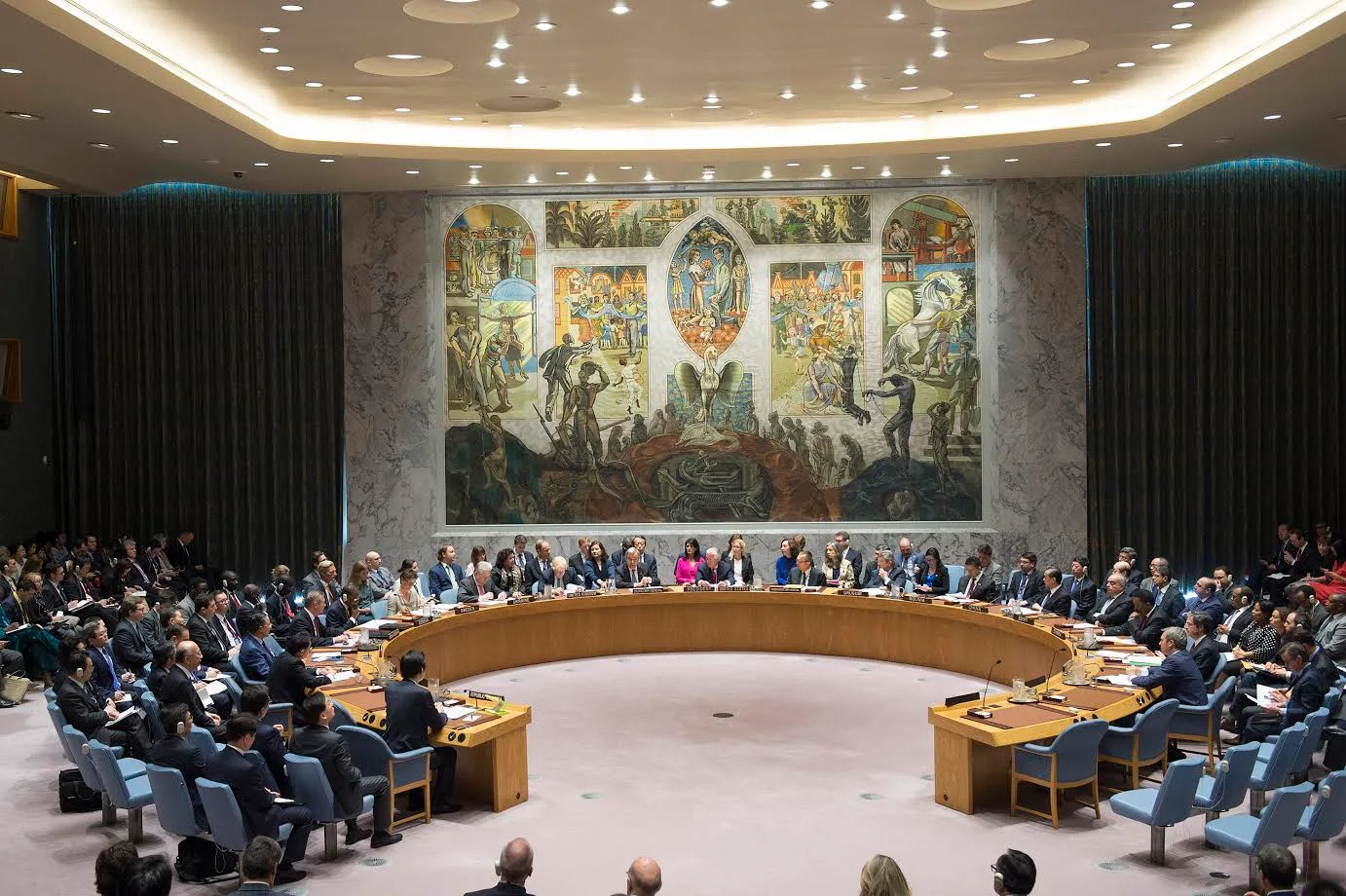 Delegates seated around the horseshoe table of the UN Security Council chamber below a large mural
