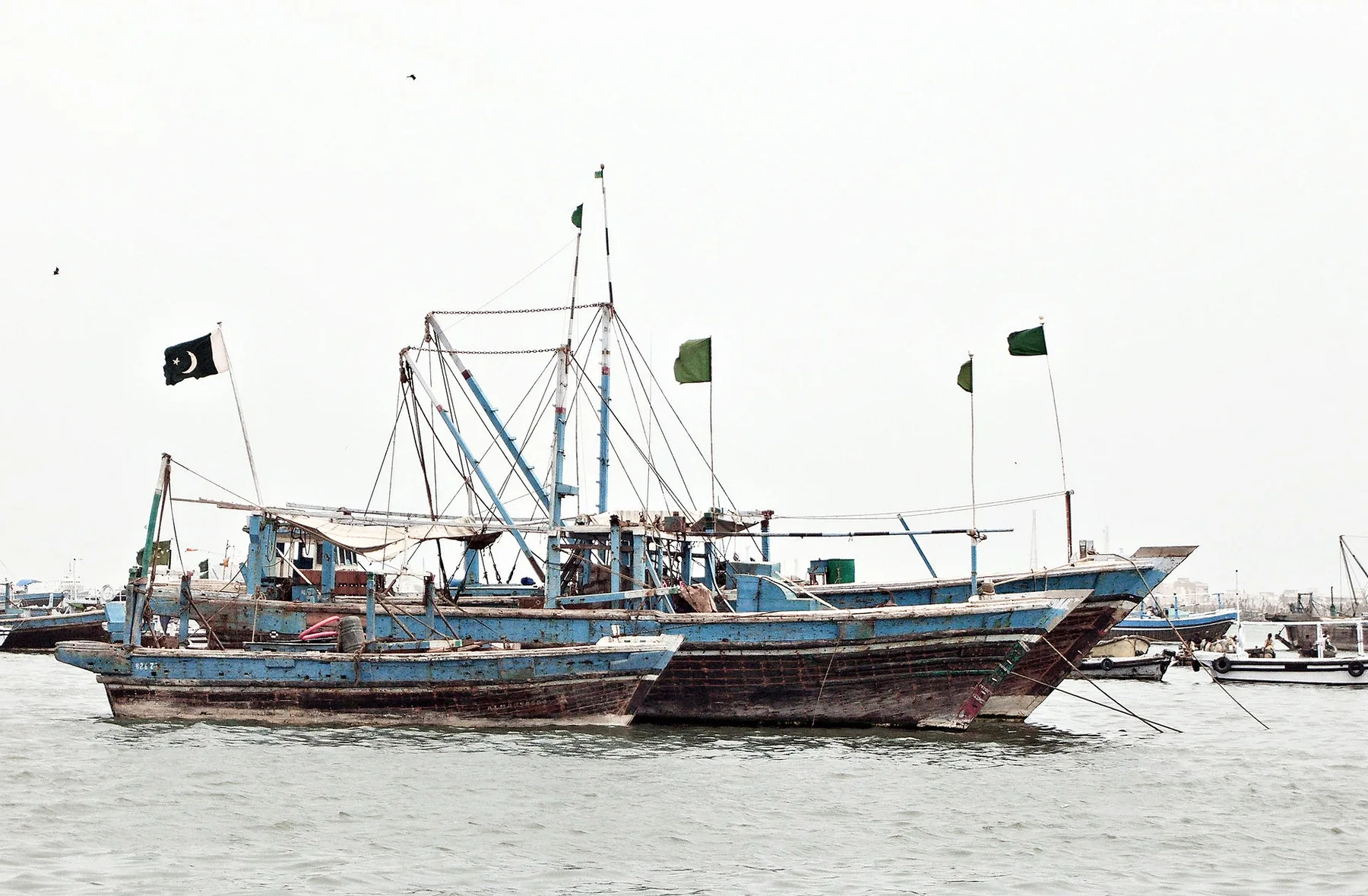 Blue wooden fishing trawlers flying Pakistani flags moored in harbour water under a grey sky