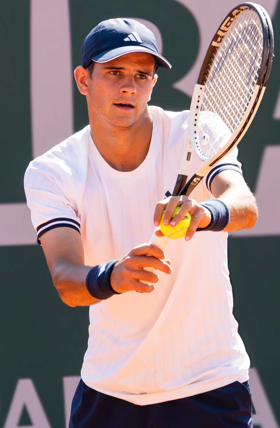Tennis player in a white shirt and navy cap holds a ball against his racket before serving