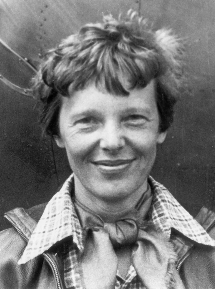 Black and white portrait of a smiling Amelia Earhart in a checked shirt and scarf beside her aircraft