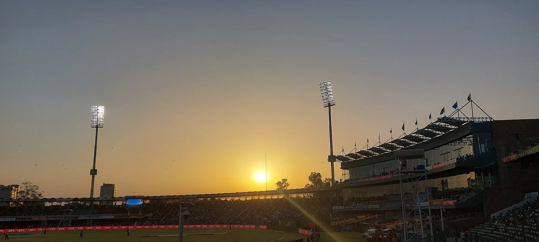 Gaddafi Stadium in Lahore at sunset with floodlights on and flags along the pavilion roof