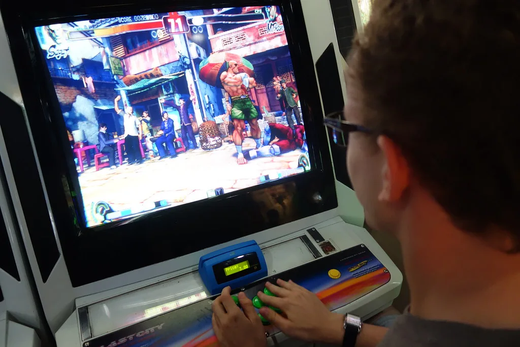 A player at an arcade cabinet presses buttons during a Street Fighter game, a wrestler character on screen