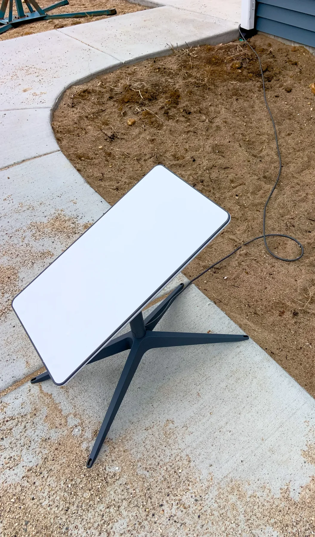 White rectangular Starlink satellite dish on a grey stand, cable running across a concrete path