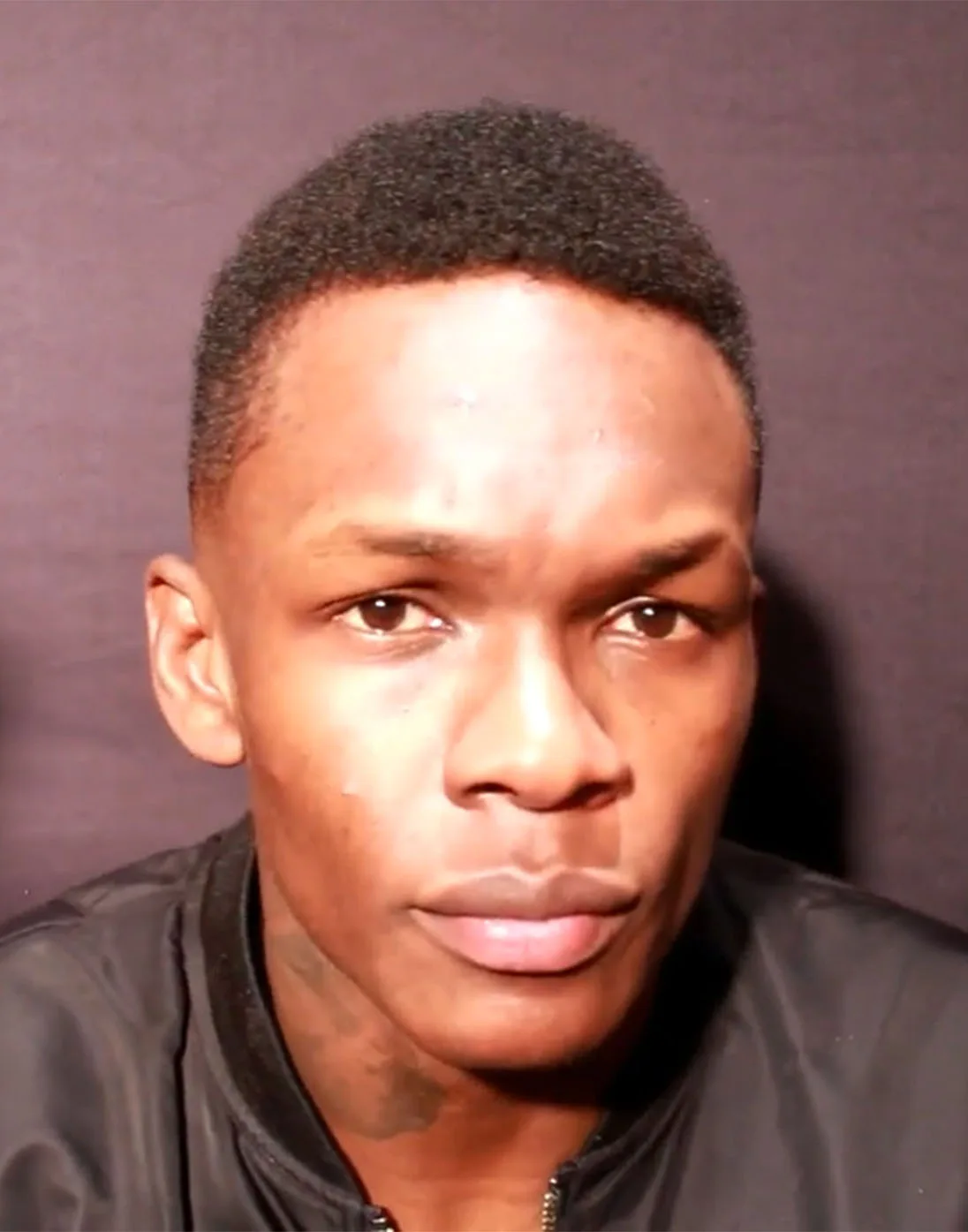 Israel Adesanya in a black jacket speaking to the camera in front of a plain backdrop