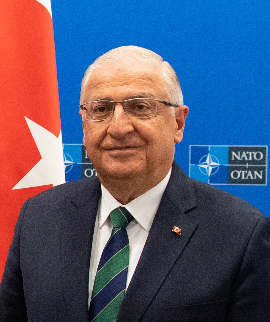 Grey-haired official in glasses and a dark suit beside a Turkish flag, NATO logo behind him