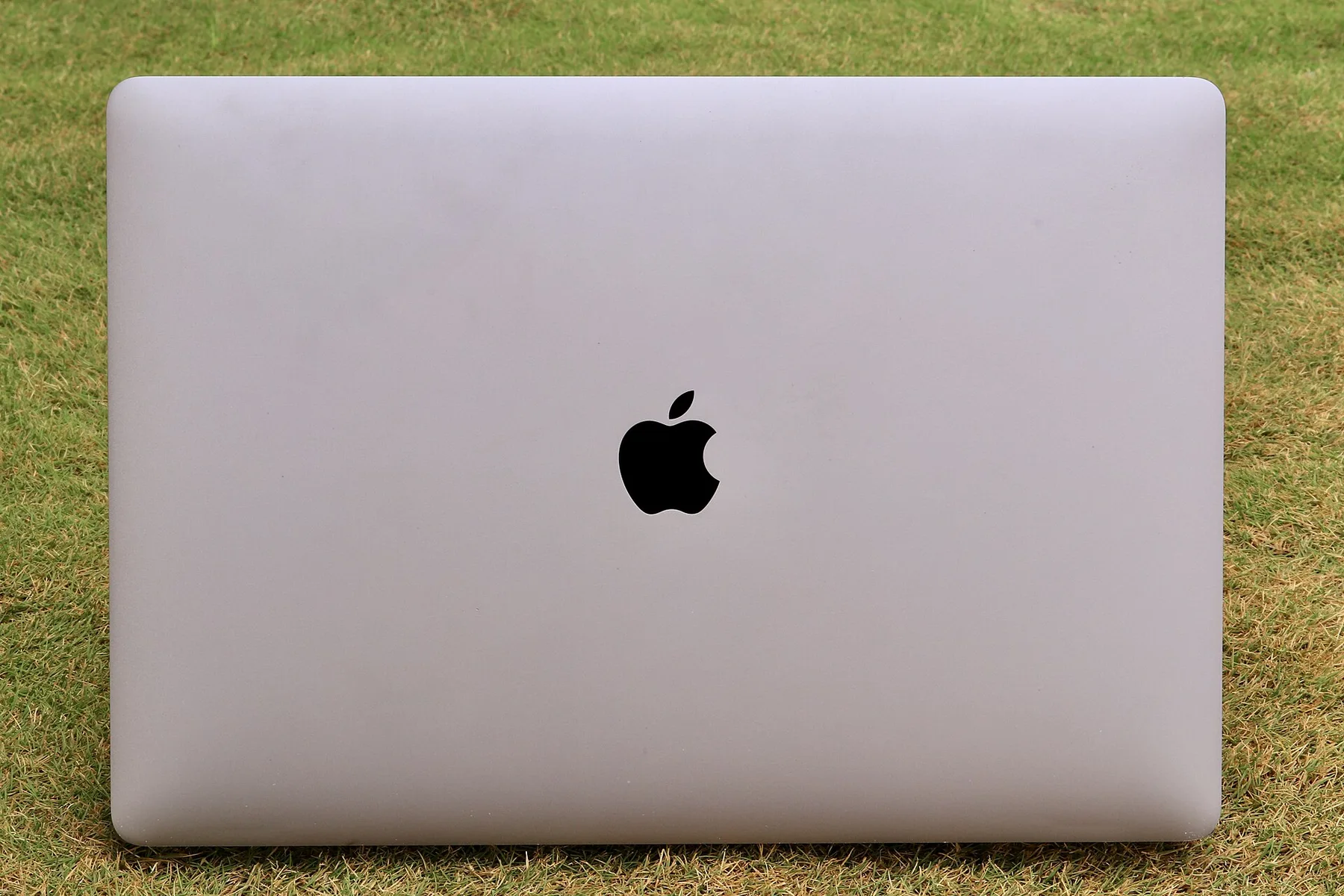 A closed silver Apple MacBook laptop with the black Apple logo, lying on green grass