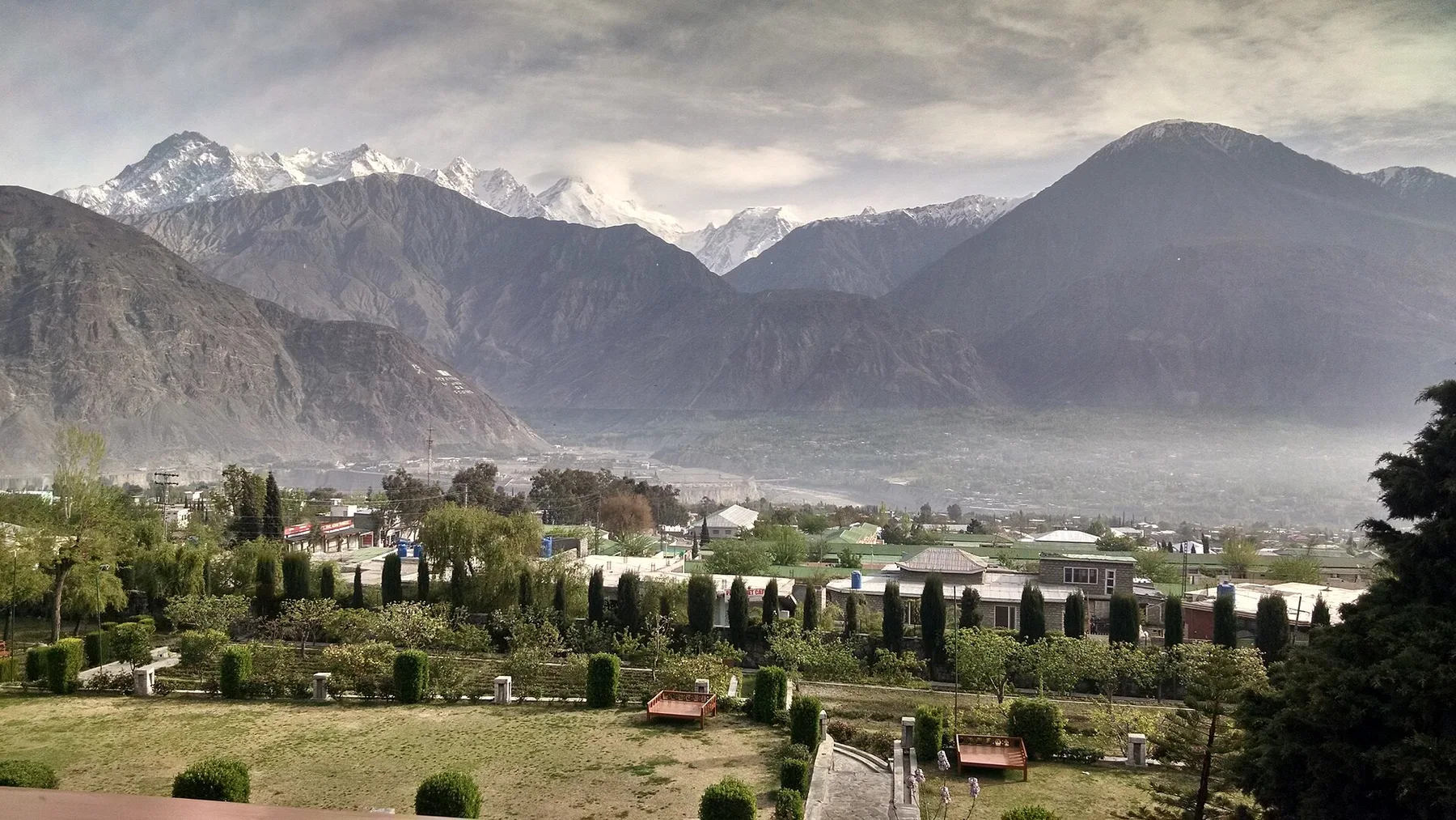 Gilgit town and green gardens below snow-capped Karakoram peaks under a cloudy sky
