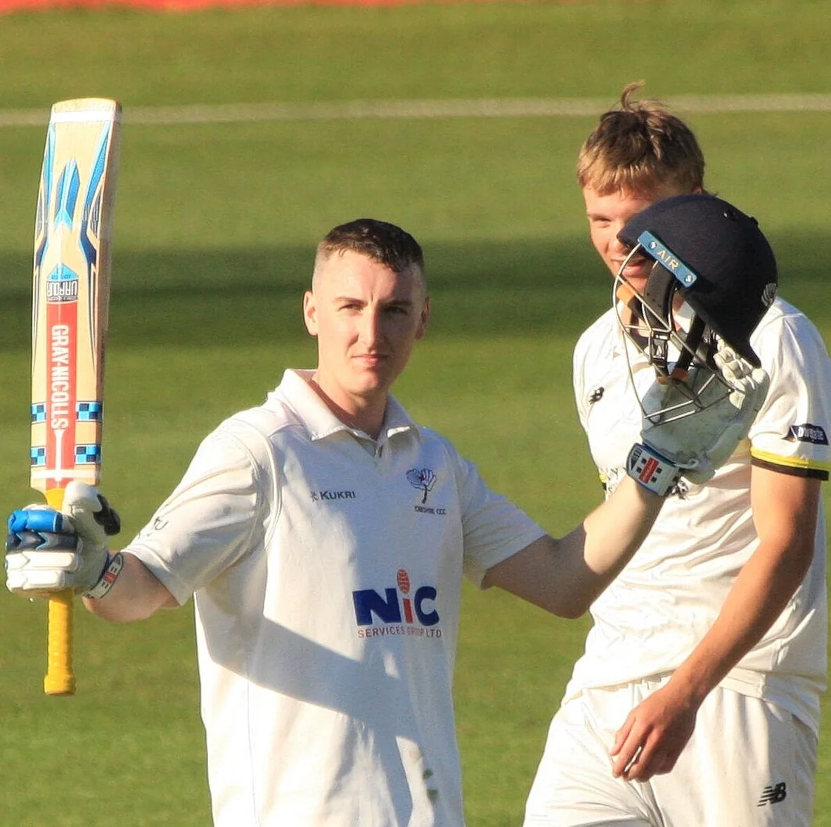 Harry Brook in Yorkshire whites raises his bat to the crowd as a teammate in a helmet looks on