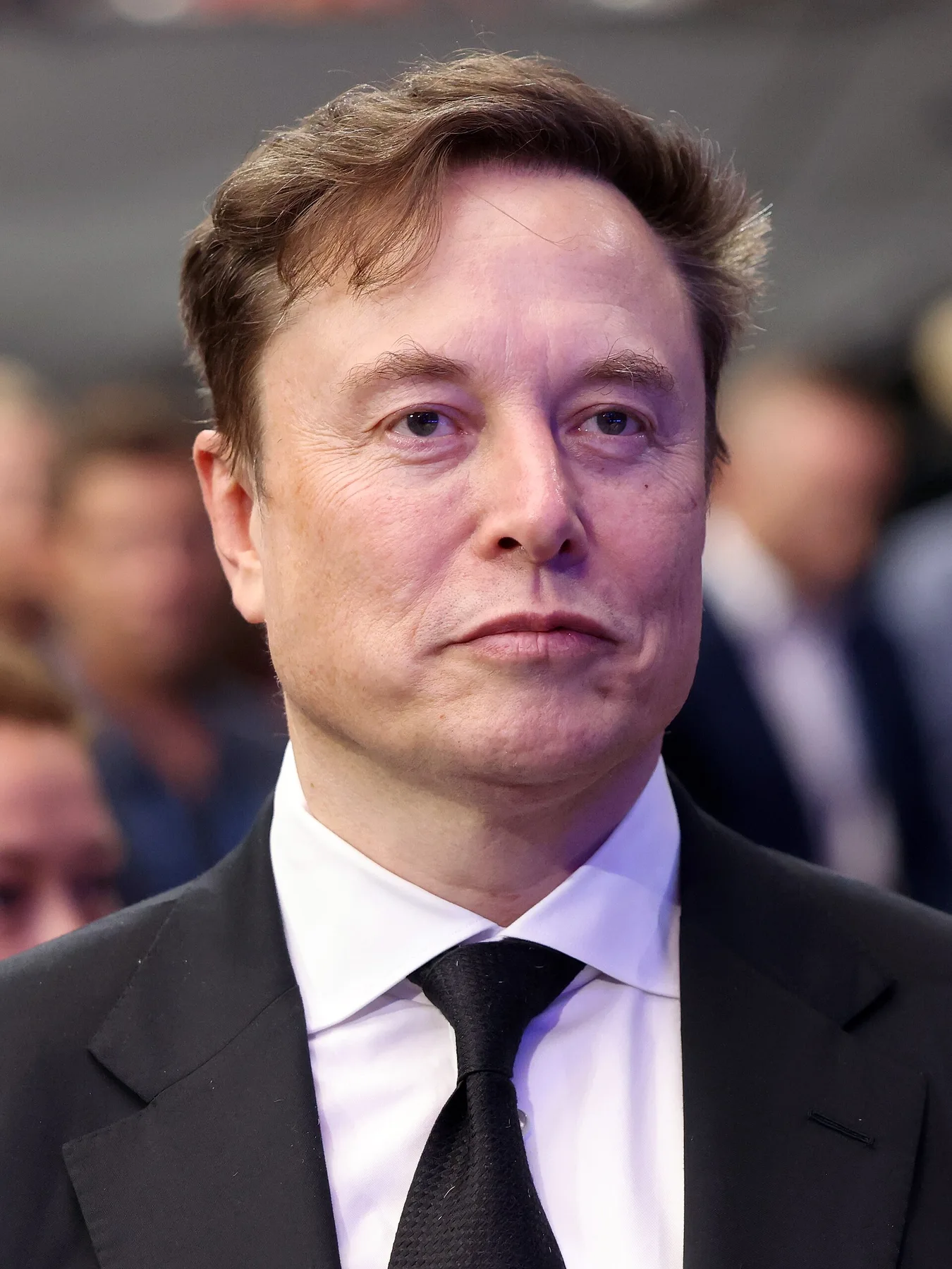 Elon Musk in a dark suit and black tie at an indoor event