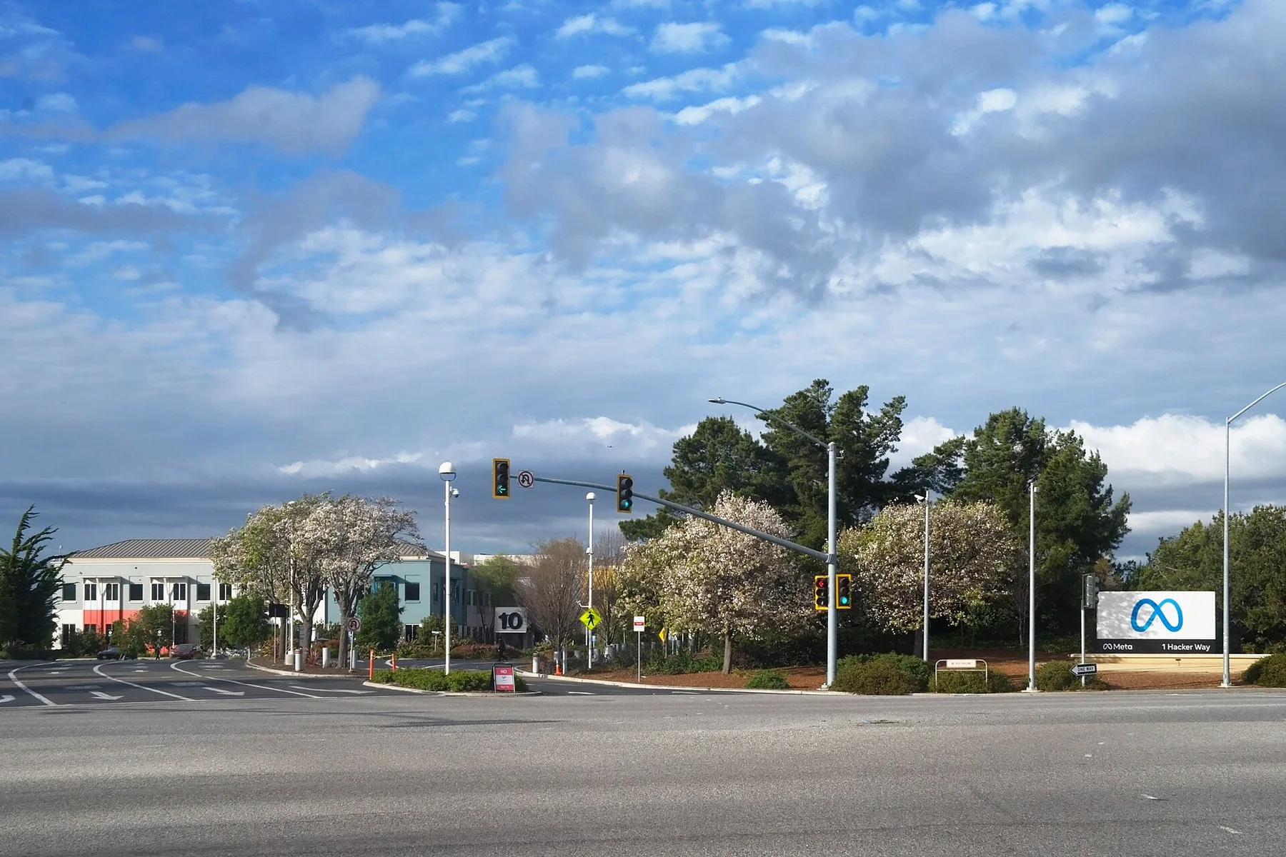 Meta sign at 1 Hacker Way beside an intersection with traffic lights and blossoming trees