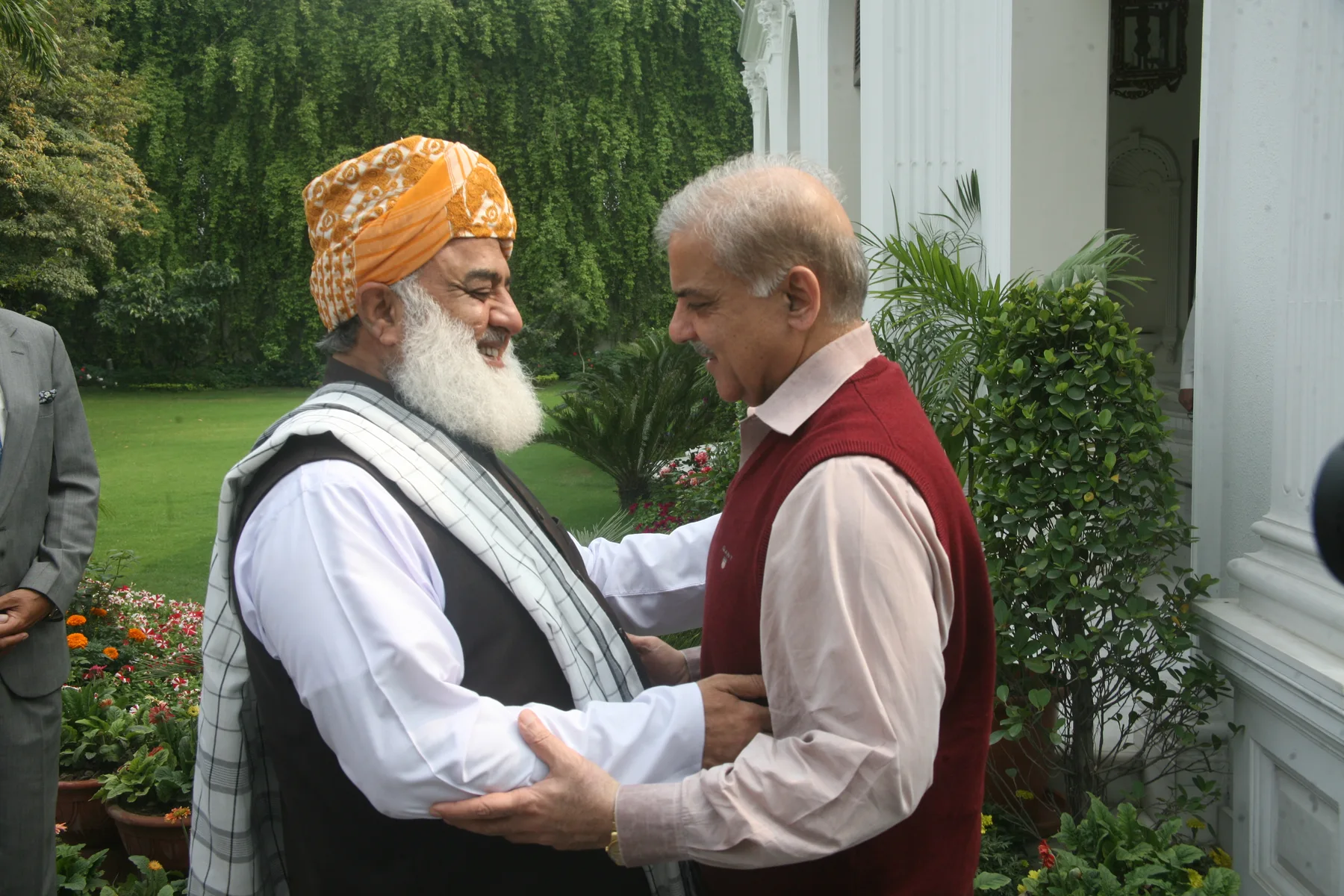 Maulana Fazlur Rehman in an orange turban embracing Shehbaz Sharif in a garden outside a white building