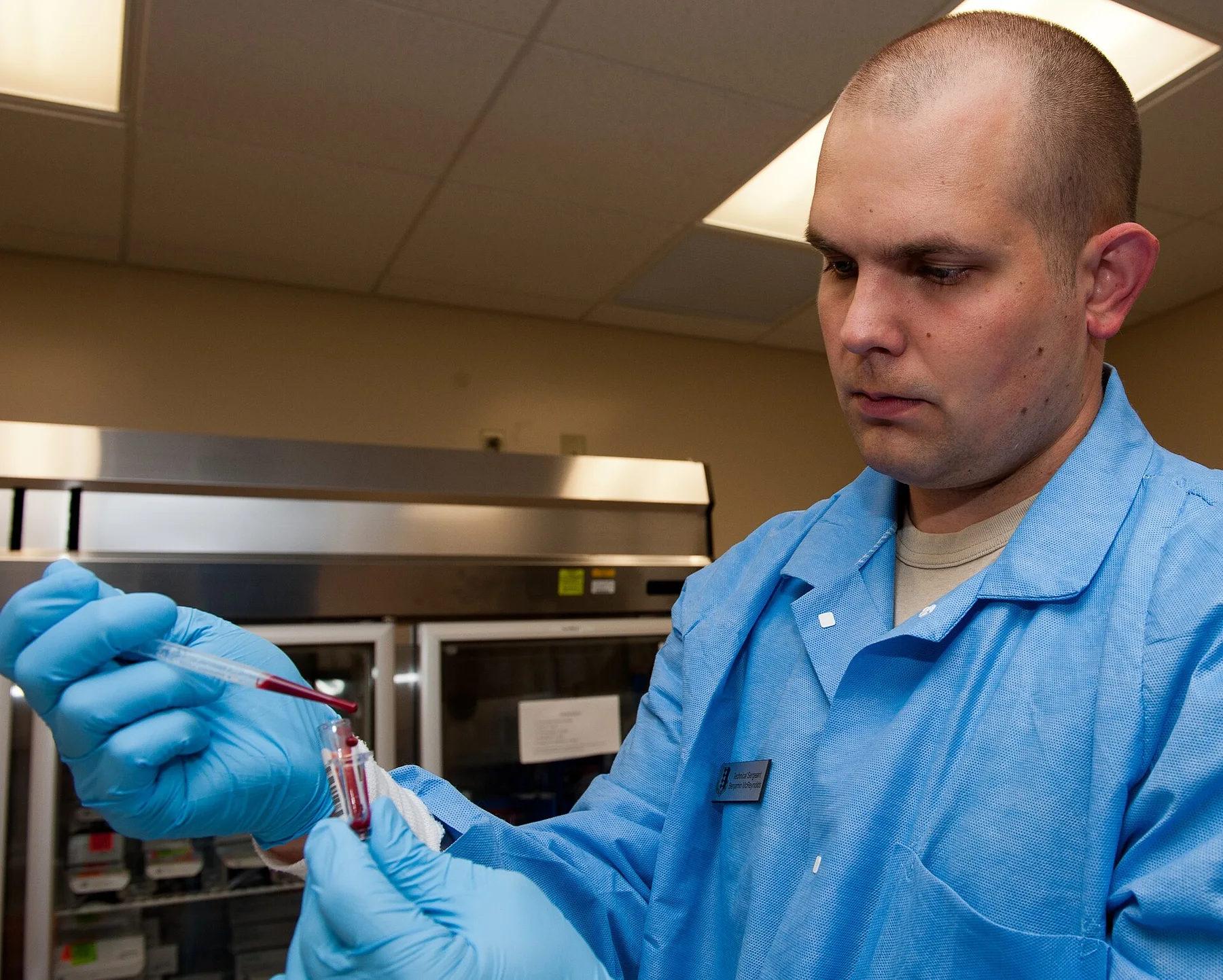 Laboratory technician in blue gloves and gown pipetting a blood sample into a test tube