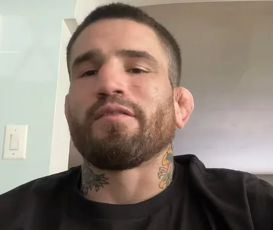 Sean Brady, bearded with neck tattoos, wearing a black T-shirt in a room at home