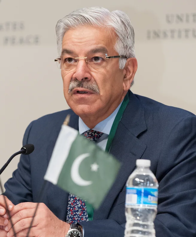 Khawaja Asif in glasses and a dark suit speaking at a conference table behind a small Pakistan flag