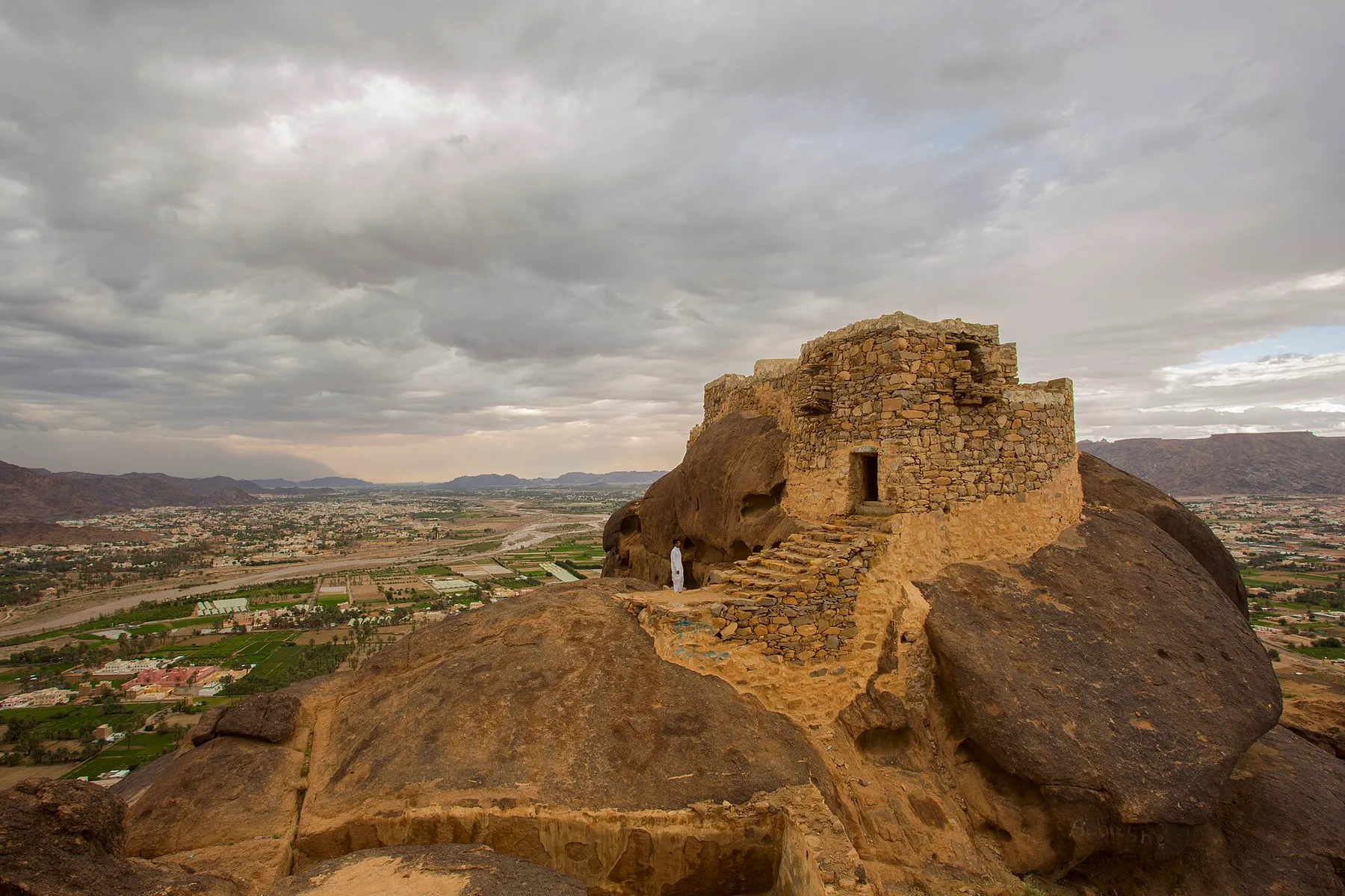 Old stone fort on a rocky outcrop above green farmland and hills in Najran province