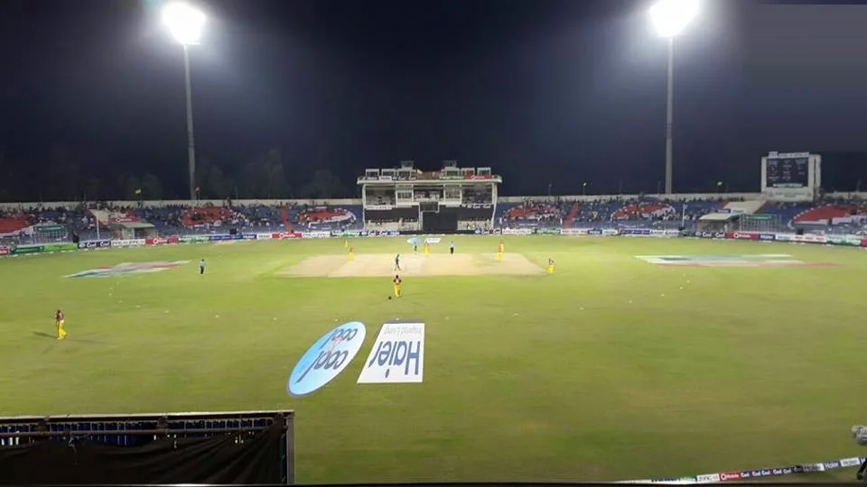 Iqbal Stadium in Faisalabad under floodlights during a night match, with players on the field