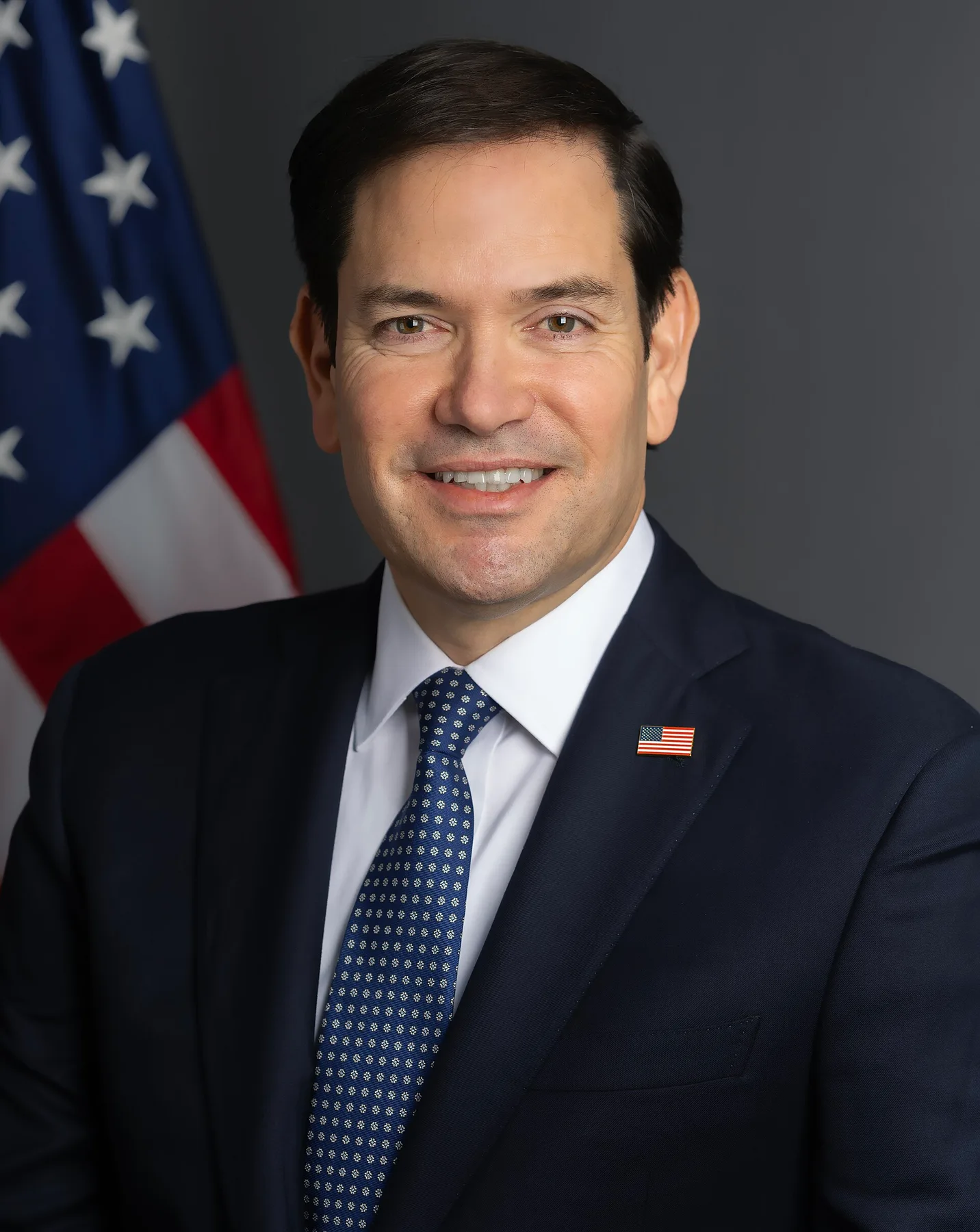 Marco Rubio smiling in a navy suit and blue tie in front of a US flag, official portrait