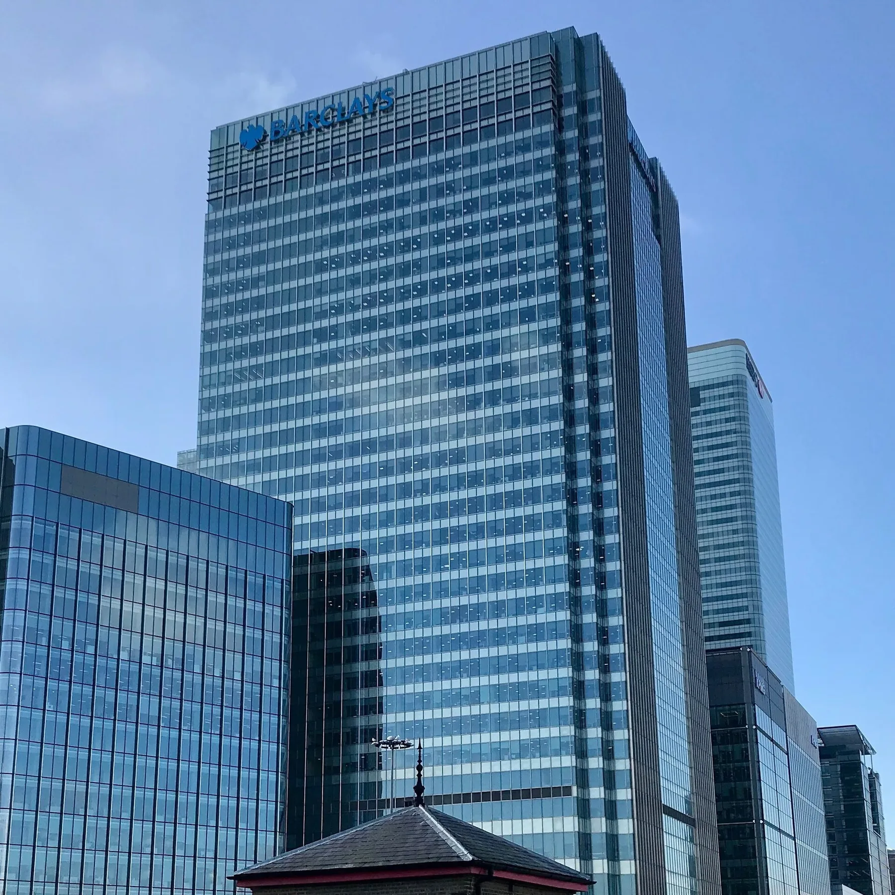 Barclays glass tower with the blue Barclays sign at the top, among office blocks in Canary Wharf