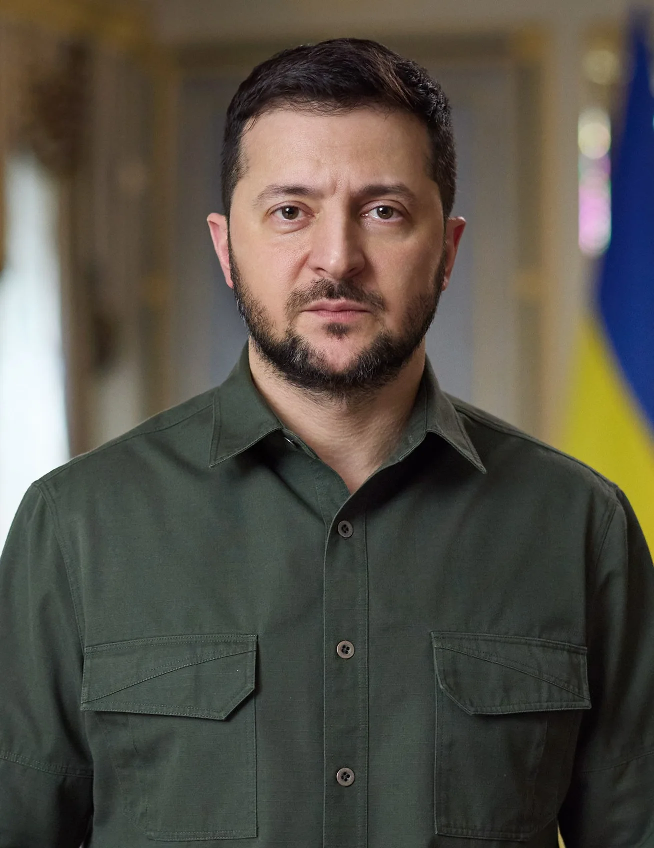 Volodymyr Zelensky in an olive military-style shirt, Ukrainian flag behind him