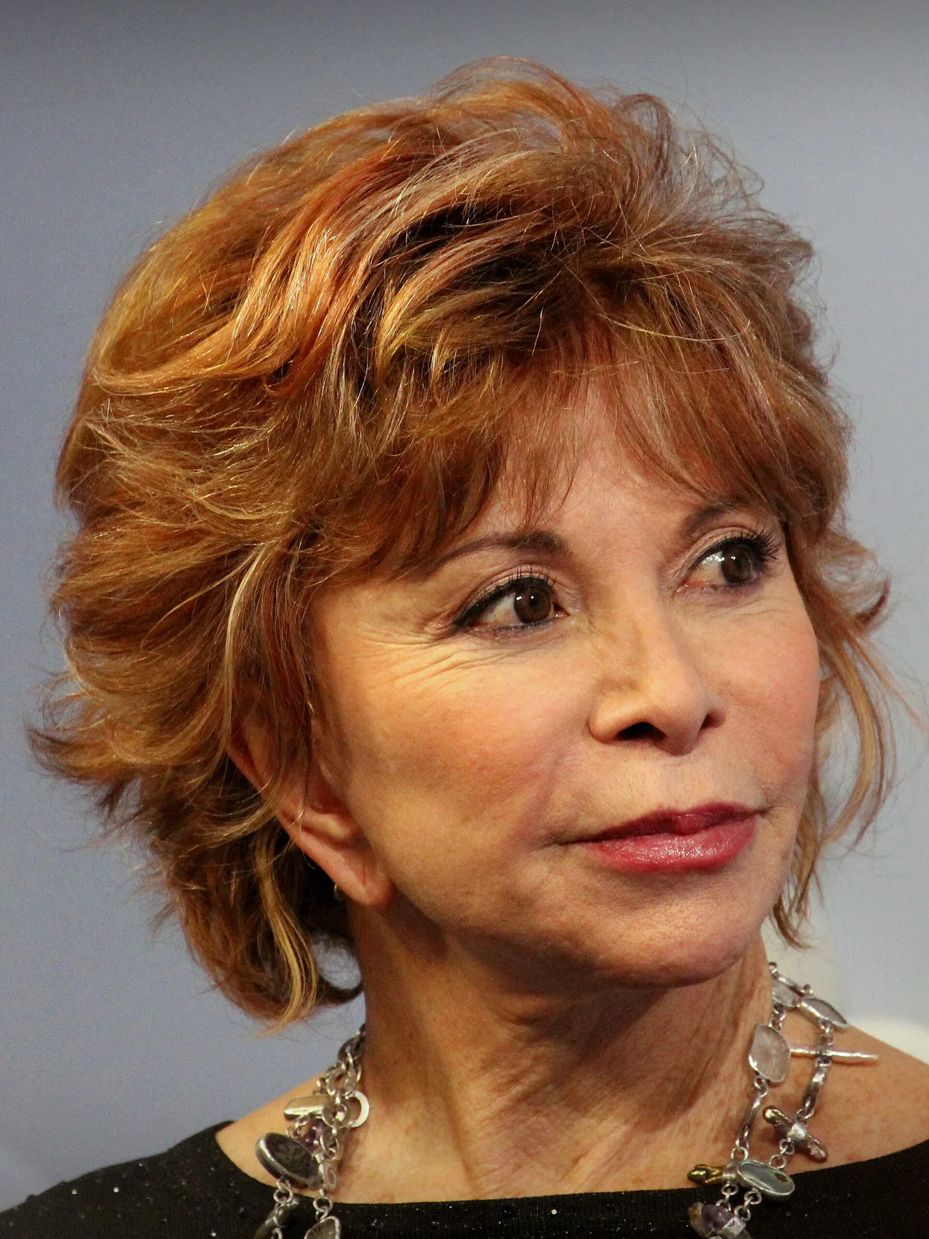 Isabel Allende with short auburn hair and a silver charm necklace, looking to one side