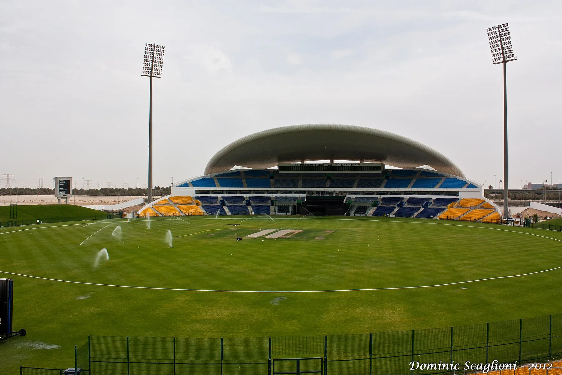 Empty Zayed Cricket Stadium in Abu Dhabi with its curved roof, blue and yellow seats and sprinklers on the outfield