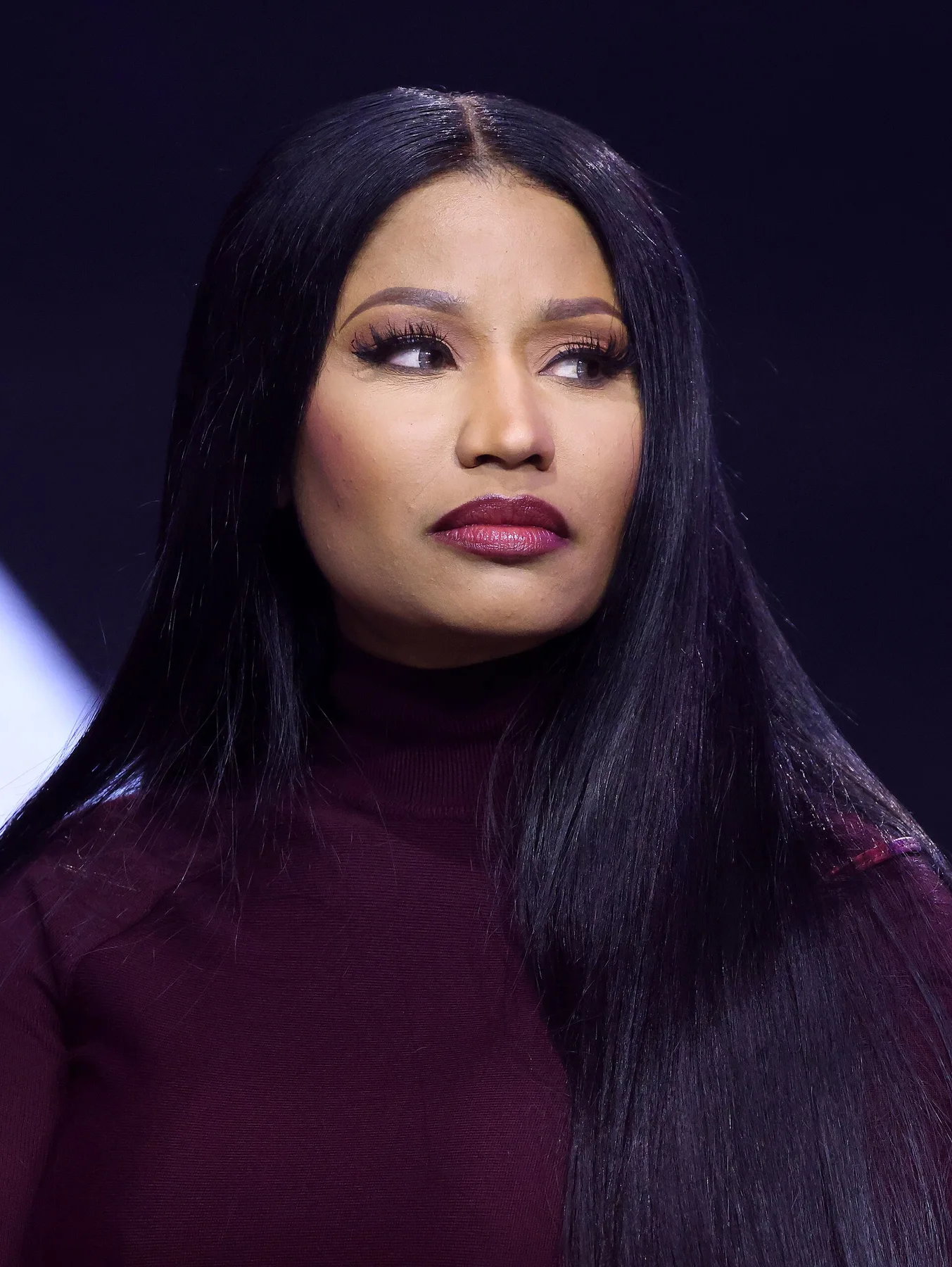 Nicki Minaj with long black hair and a maroon top, glancing to the side on a dark stage