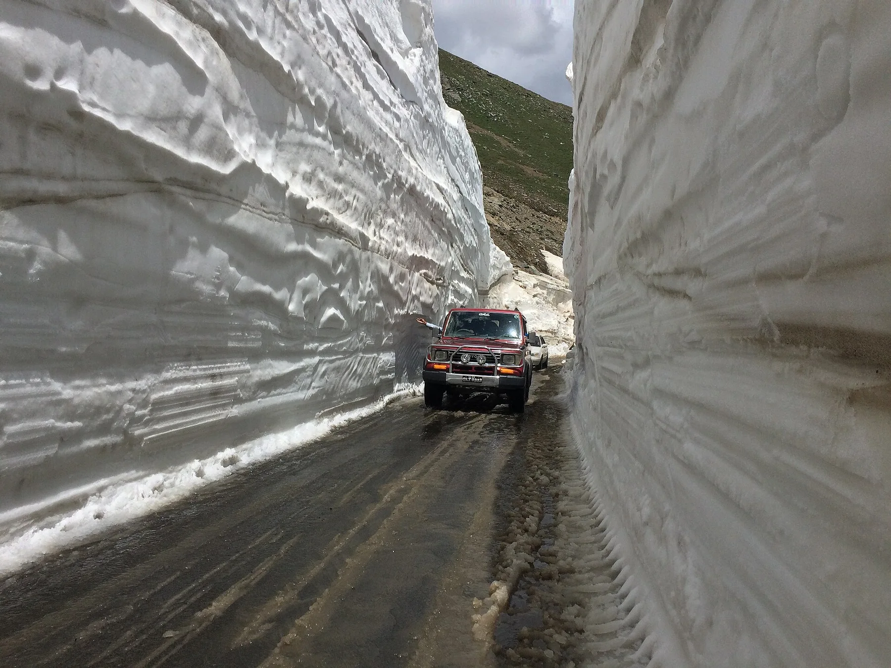 A red jeep drives along a narrow mountain road cut between towering walls of snow