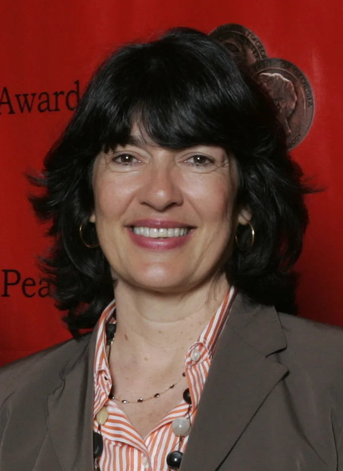 Christiane Amanpour smiling in a grey jacket and striped shirt in front of a red awards backdrop