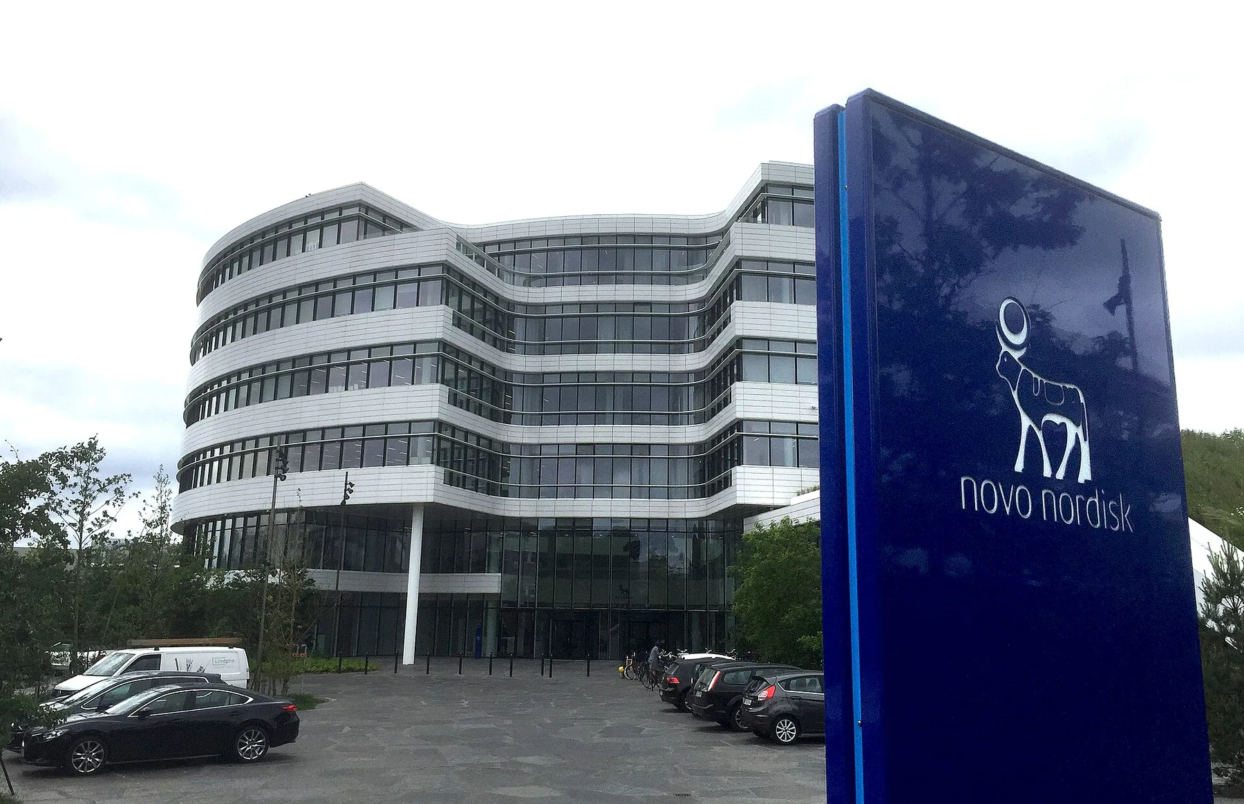 Curved glass Novo Nordisk office building behind a tall blue sign with the company logo, cars parked in front