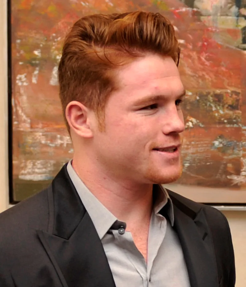 Canelo Alvarez in a dark jacket and grey shirt, smiling, in front of an abstract painting