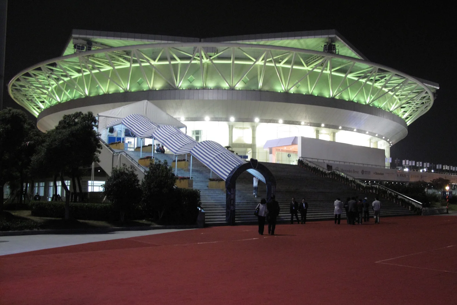 Qizhong Forest Sports City Arena in Shanghai lit green at night, visitors walking across the red forecourt