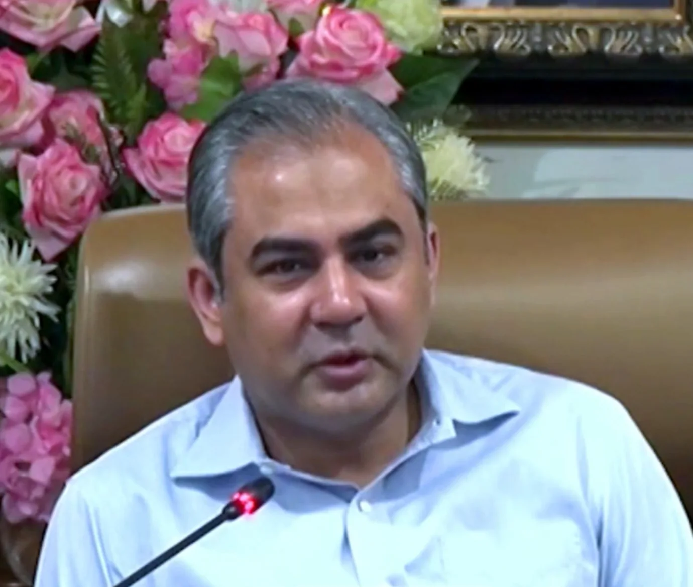 Mohsin Naqvi in a light blue shirt speaking into a microphone in front of a pink flower arrangement