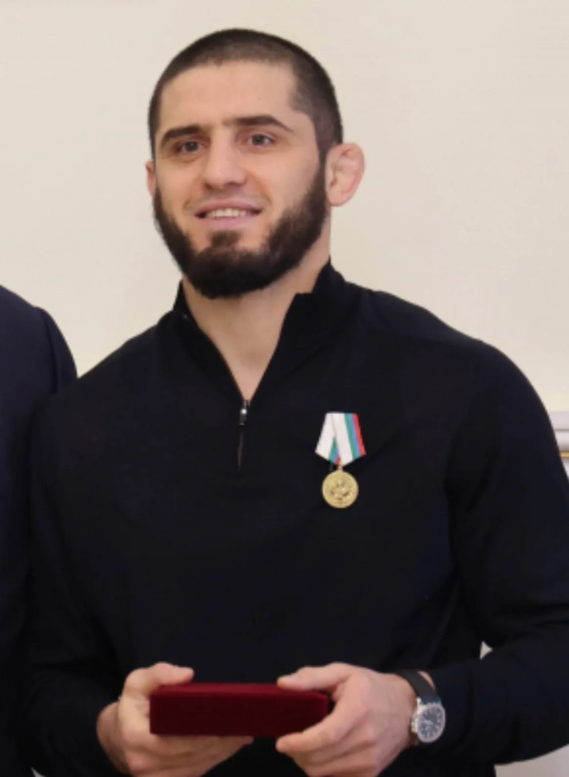 Islam Makhachev in a black zip top smiles while holding a red box, a medal pinned to his chest