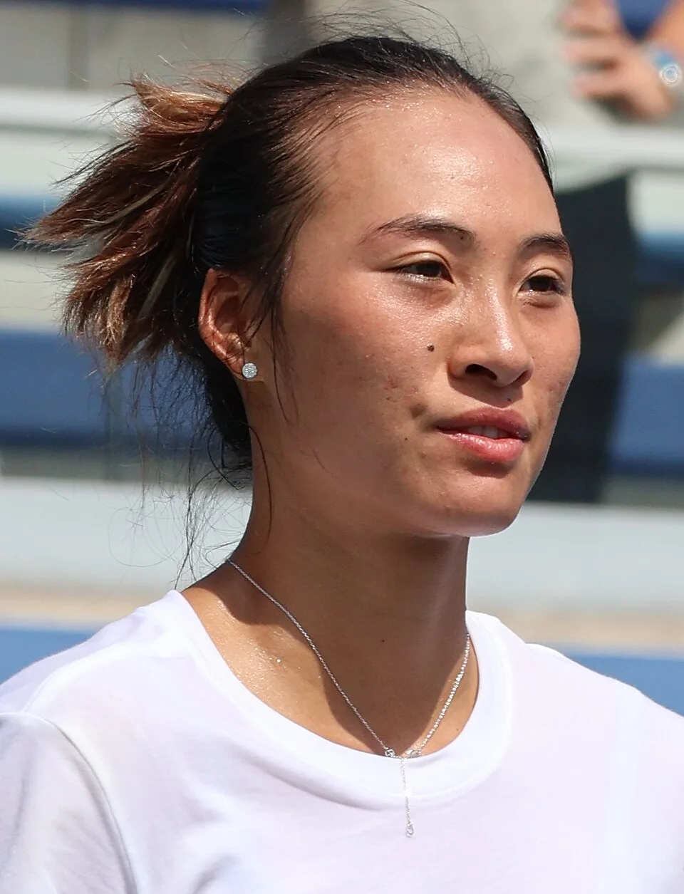 Zheng Qinwen in a white T-shirt and silver necklace at a tennis court, hair tied back