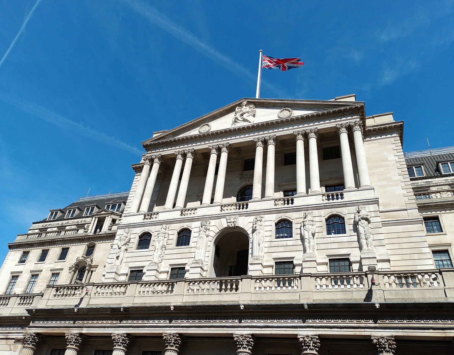 Columned stone facade of the Bank of England with the Union flag flying above against a blue sky