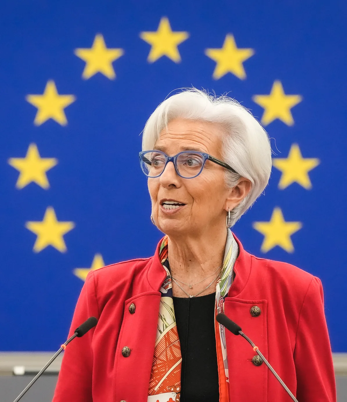 Christine Lagarde in a red jacket speaking at a lectern in front of the EU flag