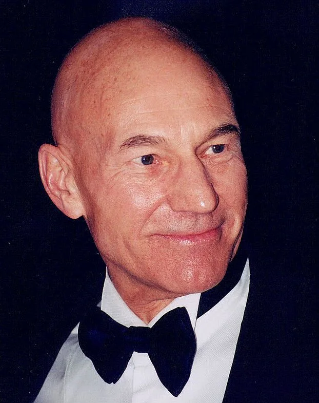 Patrick Stewart smiling in a black tuxedo and bow tie against a dark background