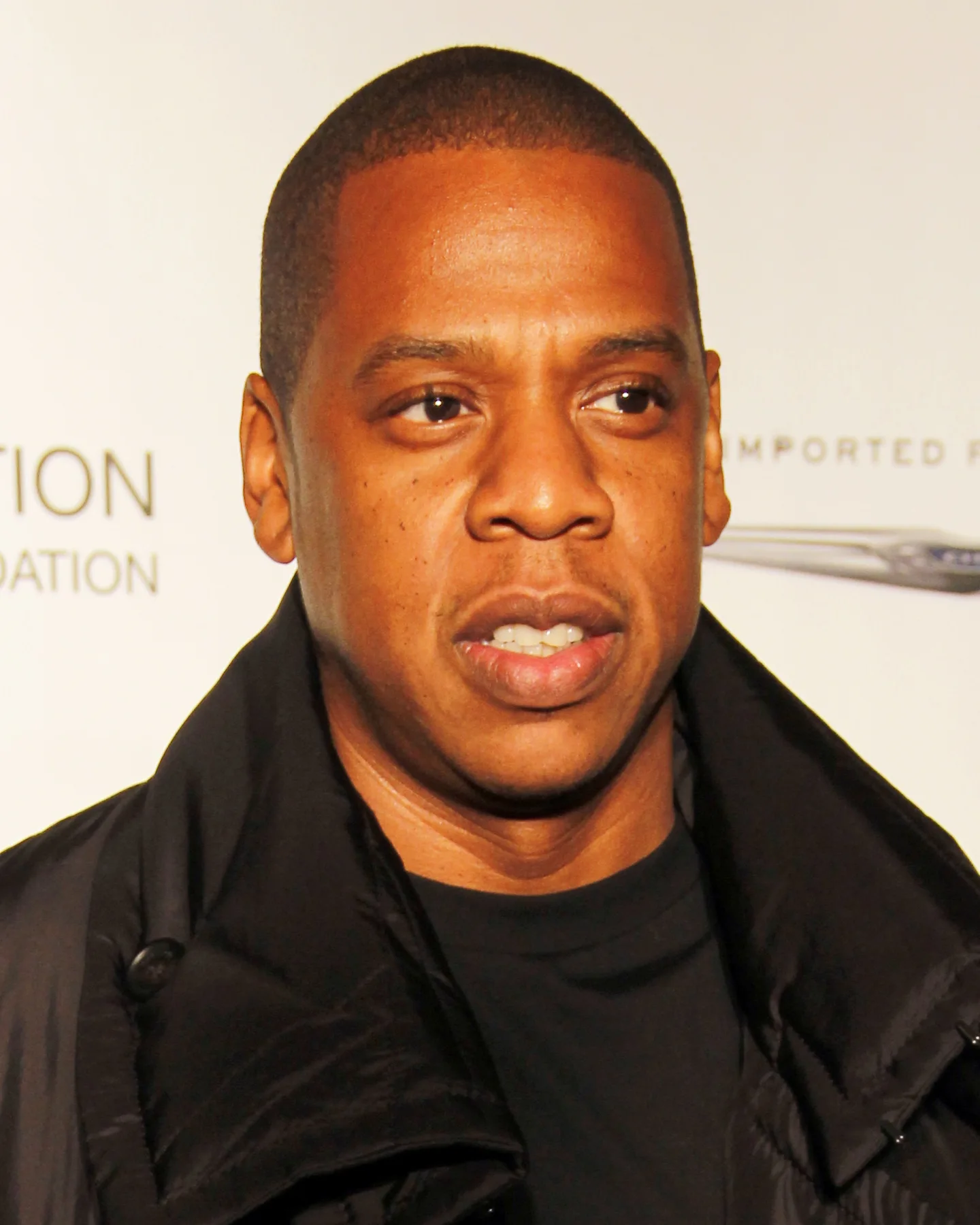 Jay-Z in a black jacket at a foundation event, close-up against a white sponsor backdrop