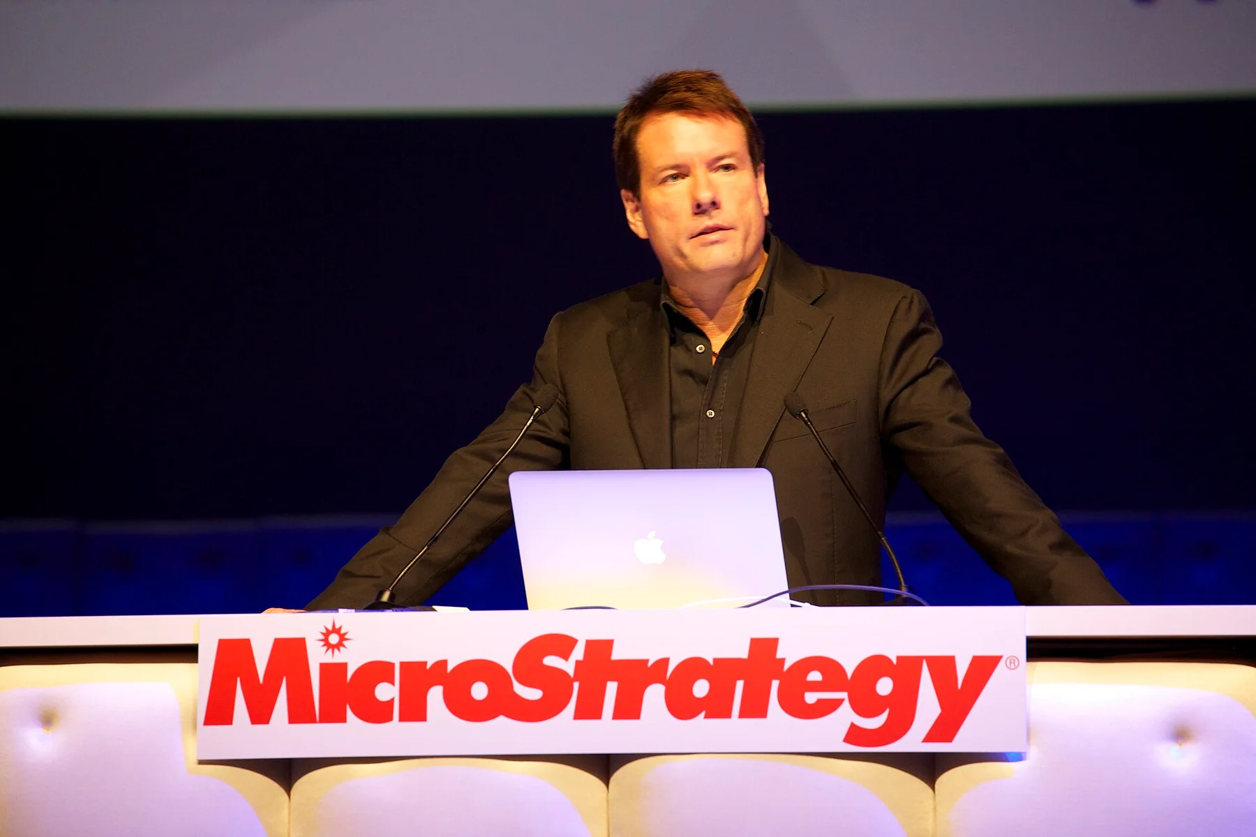 Speaker at a podium with a laptop behind a red MicroStrategy sign at a company conference