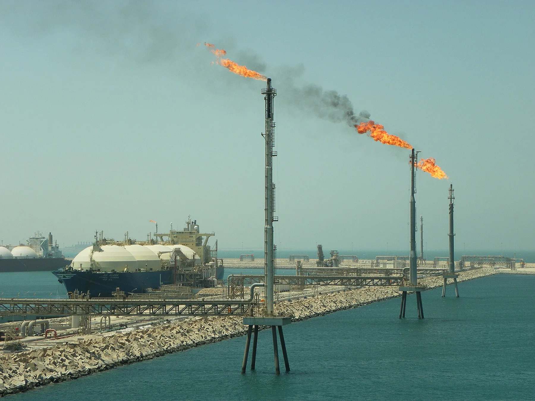 LNG tanker with white dome tanks berthed at a gas terminal jetty as flare stacks burn over the sea