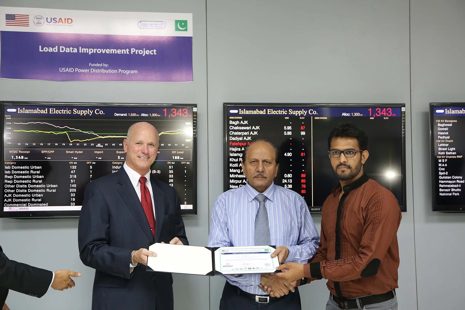 Officials hold a certificate in front of Islamabad Electric Supply Co. load monitoring screens