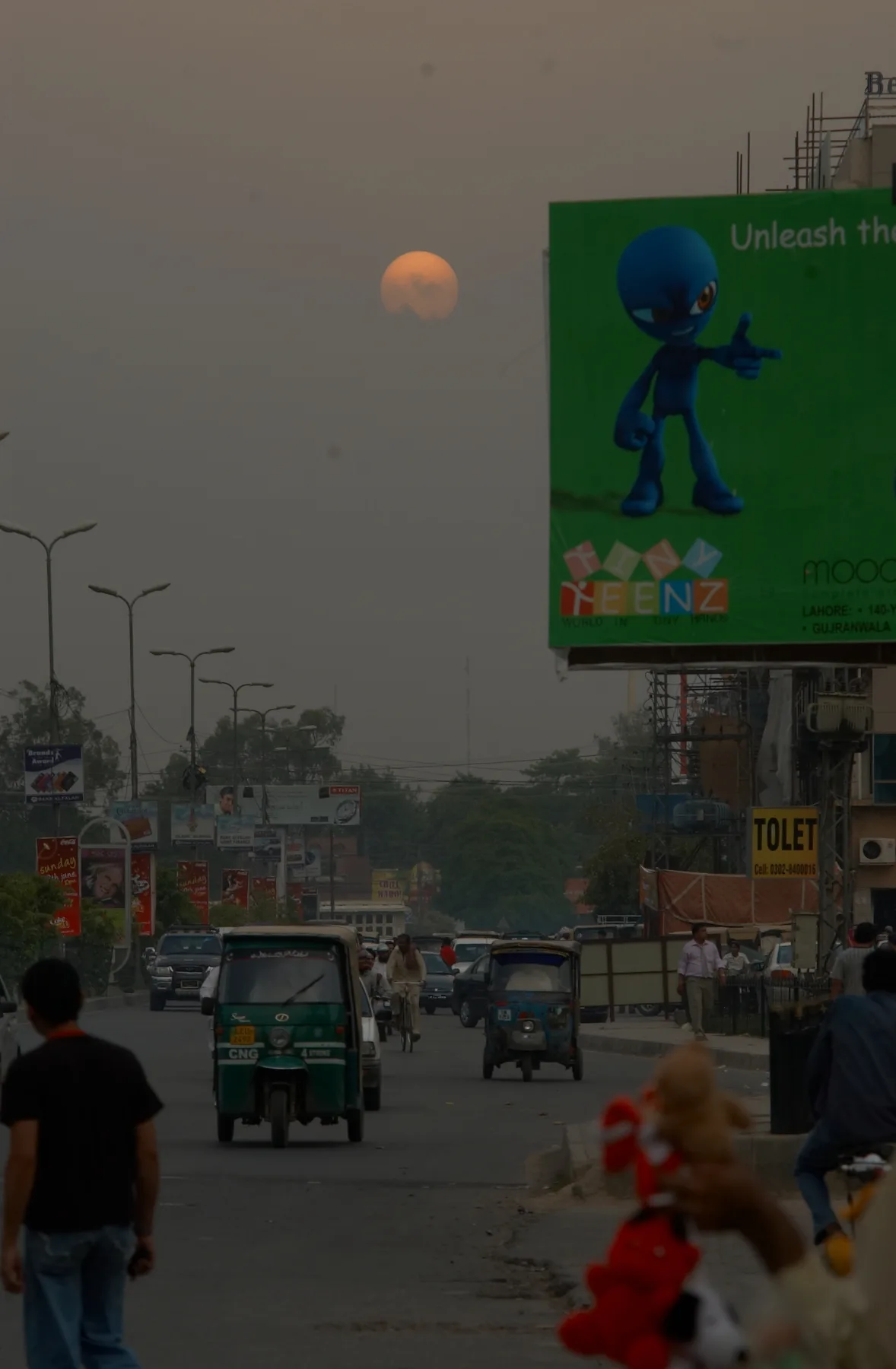 Hazy orange sun over a smoggy Lahore road with rickshaws, traffic and billboards