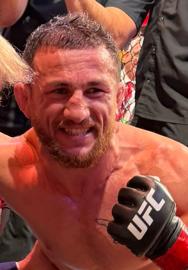 Merab Dvalishvili smiling in the octagon after a fight, raising a fist in a UFC glove