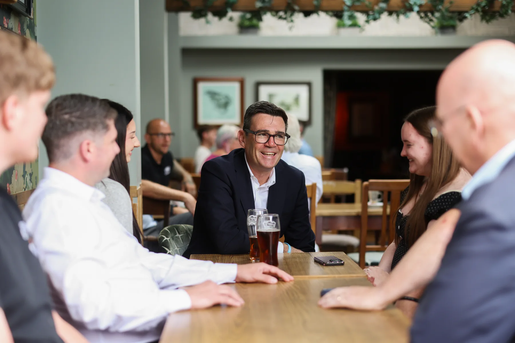 Andy Burnham in glasses and a dark jacket smiling at a pub table with a group of people