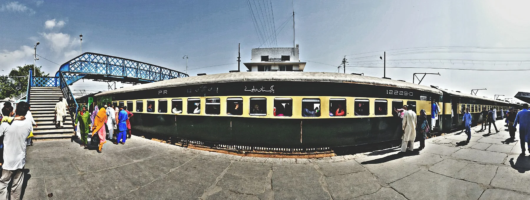Green and cream Pakistan Railways passenger coach at a station platform, passengers walking beside it under a footbridge