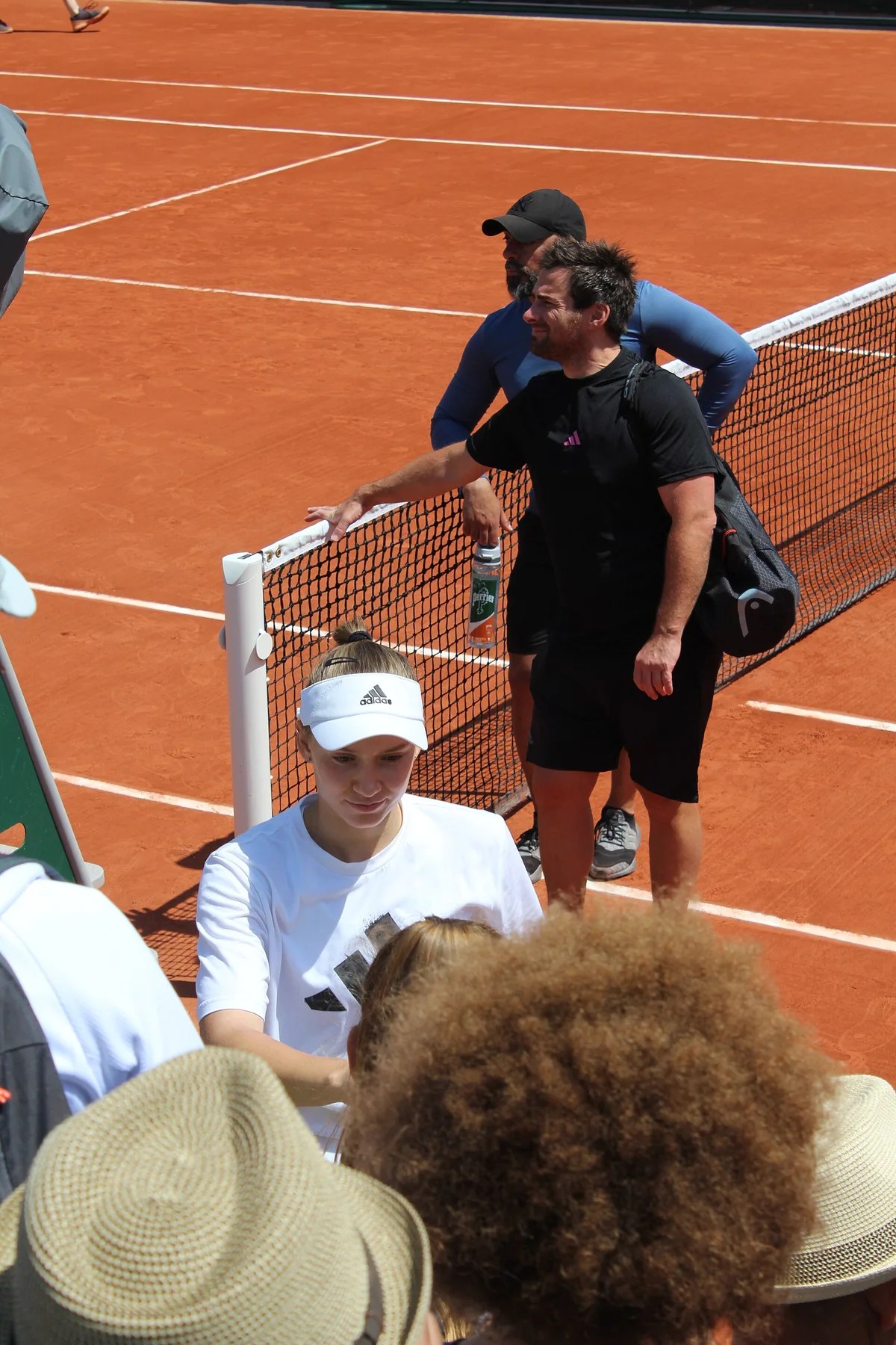 Elena Rybakina in a white visor walking off a clay court past the net as fans watch