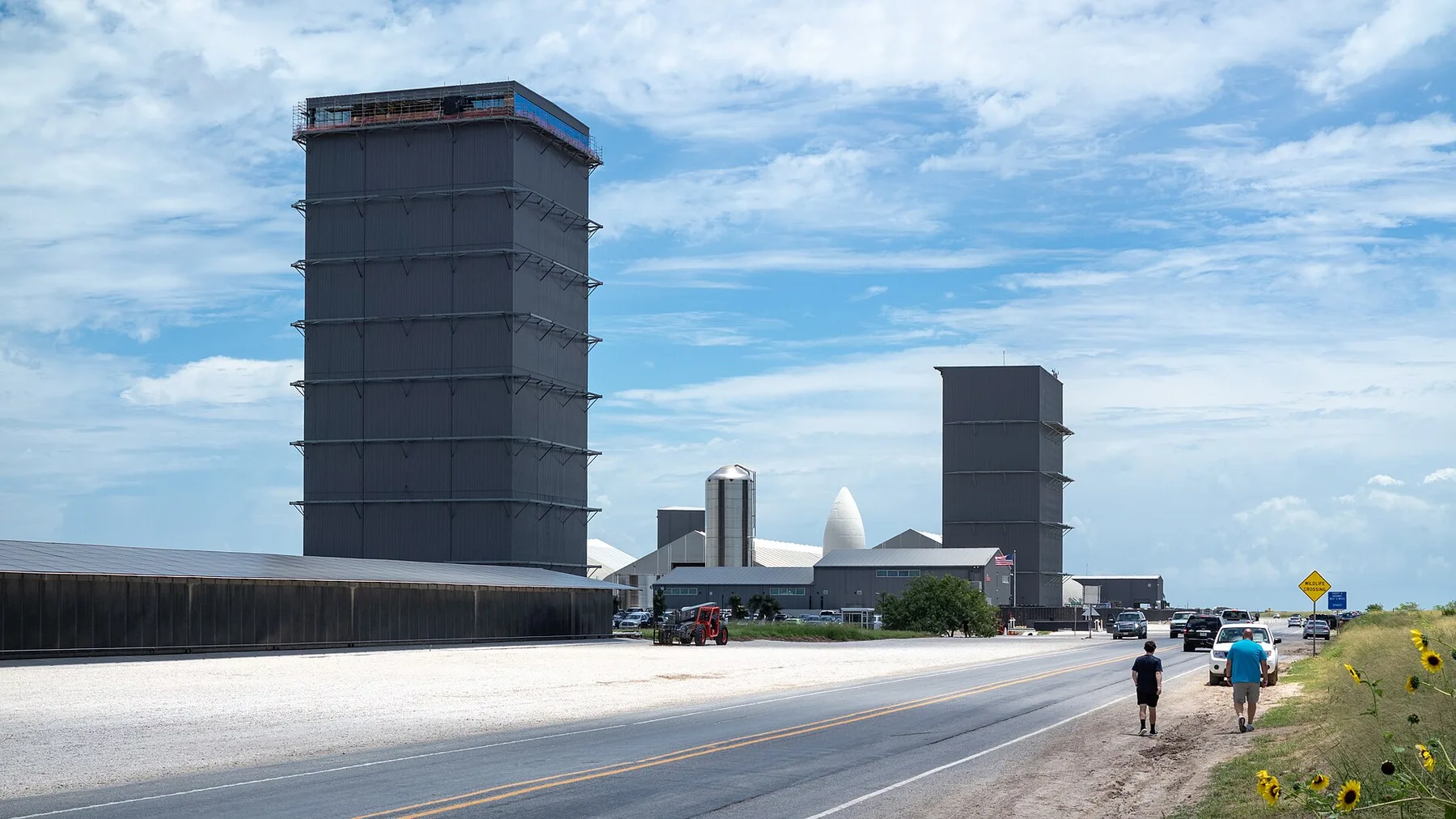 SpaceX rocket assembly towers and a white nose cone beside a road as two people walk past