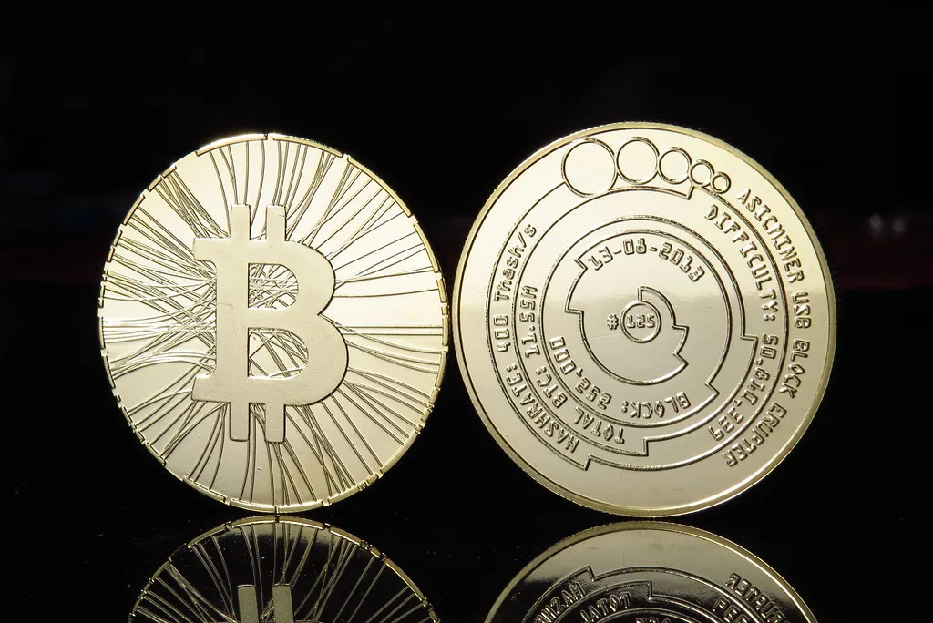 Two gold-coloured commemorative bitcoin coins, one showing the B symbol, on a black reflective surface
