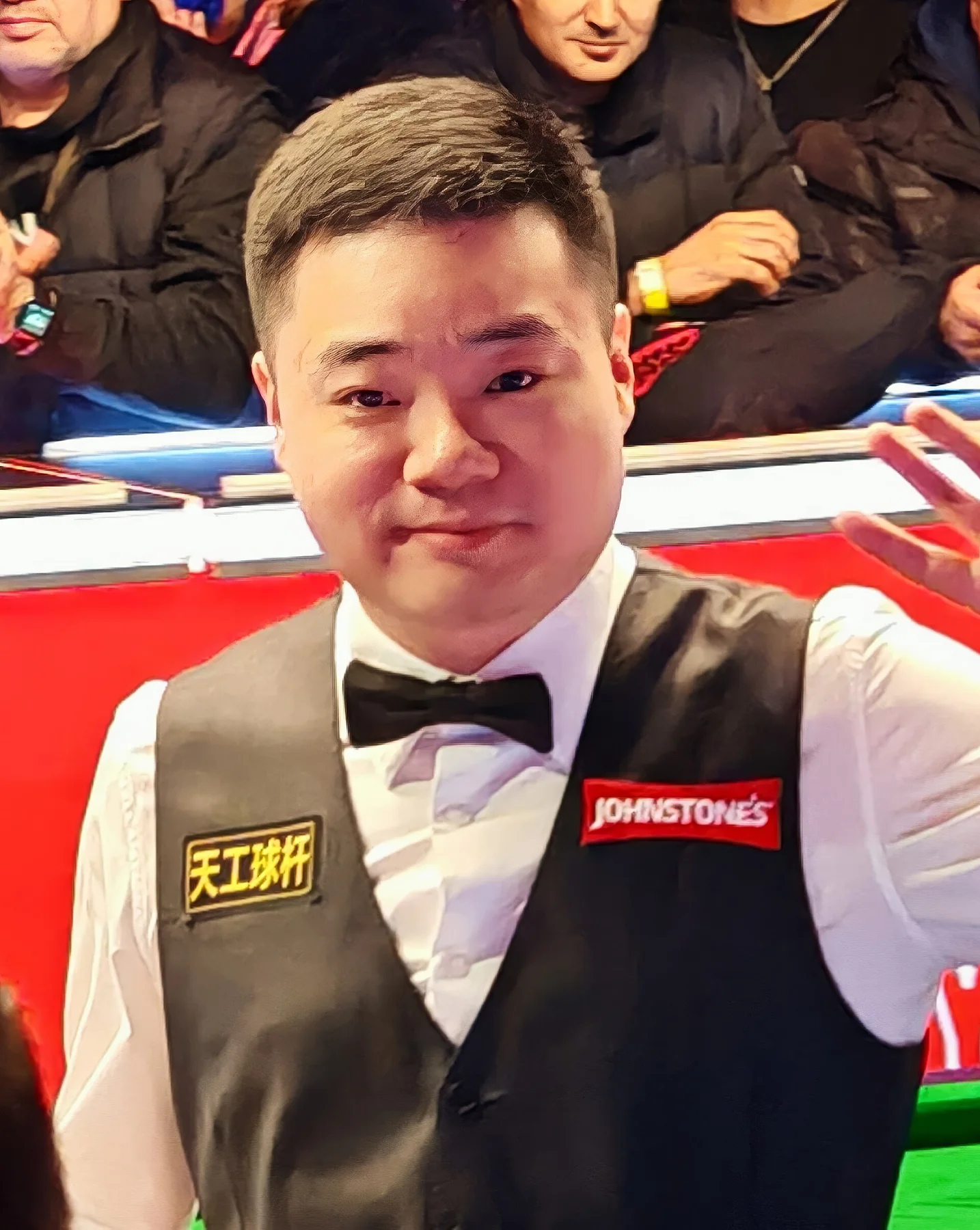Snooker player in waistcoat and bow tie waves beside the table with spectators seated behind him