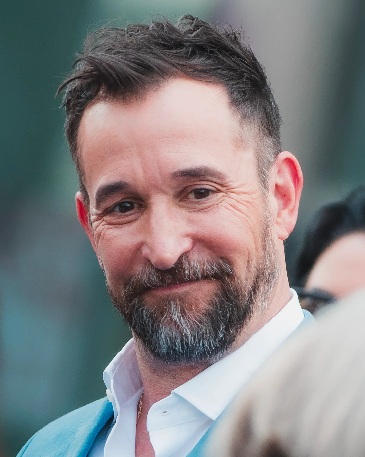 Noah Wyle, smiling with a greying beard, in a white shirt and light blue jacket at an outdoor event